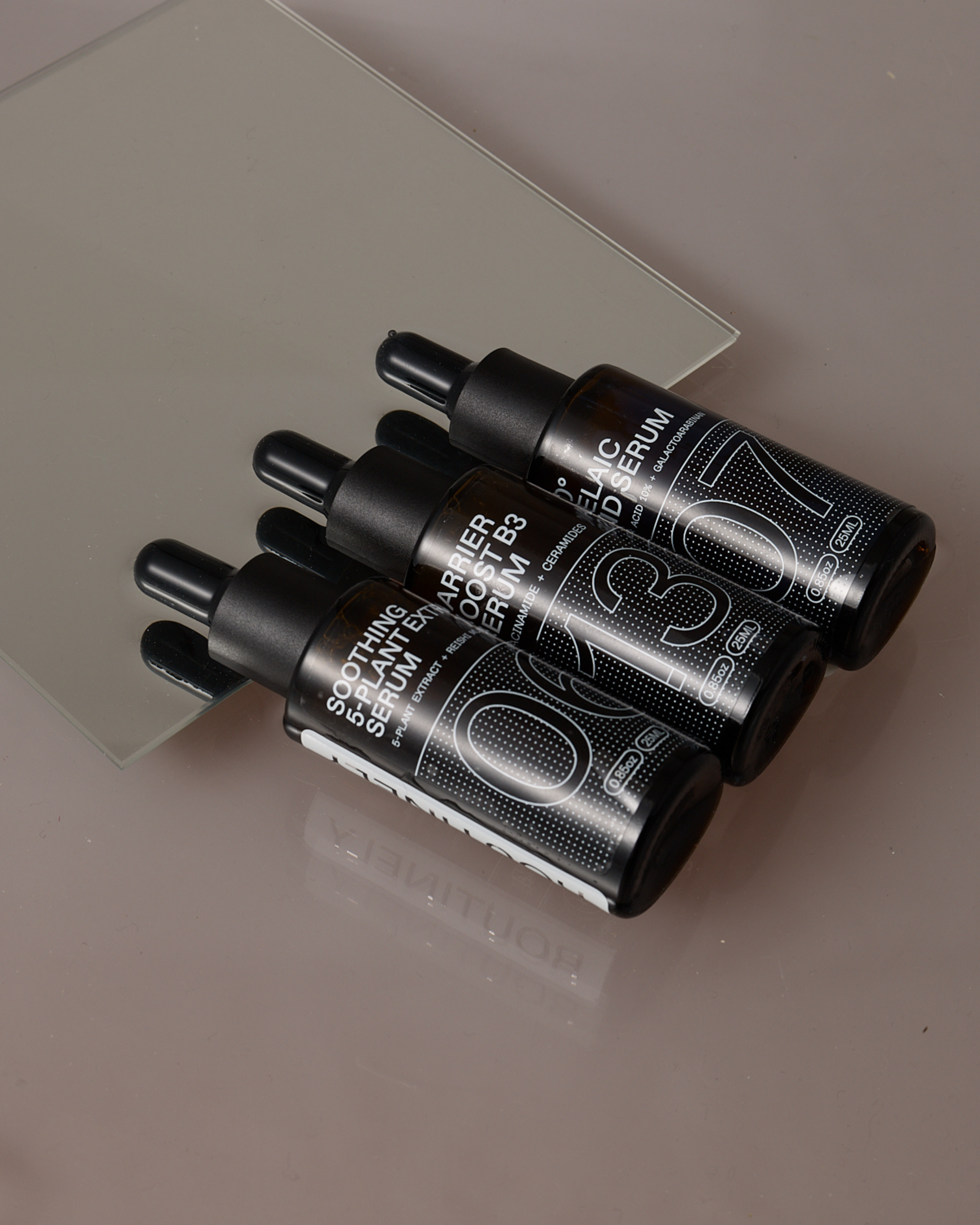 Three serums with black bottle and white logo placed in a row with a mirror background